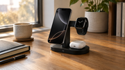 A phone, smartwatch, and earbuds sit on a charging stand on a sunlit wooden desk.