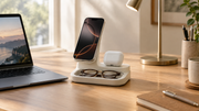 A phone, earbuds, and glasses sit on a charging stand beside a laptop on a sunlit desk.