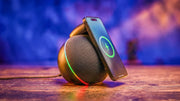 Dark grey NYTSTND ORBE Qi2.2 wireless charging speaker with an ambient light ring, charging an iPhone on a wooden table