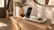 A smartphone, earbuds, and keys sit on a charging tray atop a sunlit wooden console.